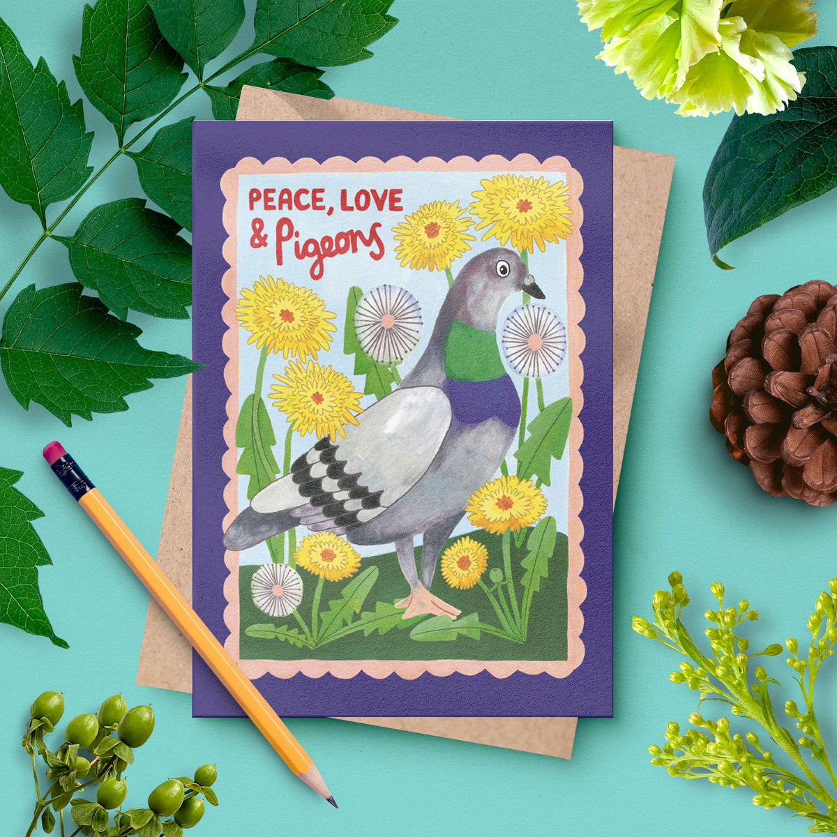 A greeting card with a purple border and an illustration of a pigeon surrounded by yellow flowers, placed on a kraft envelope with a pencil and green leaves around it on a turquoise background.